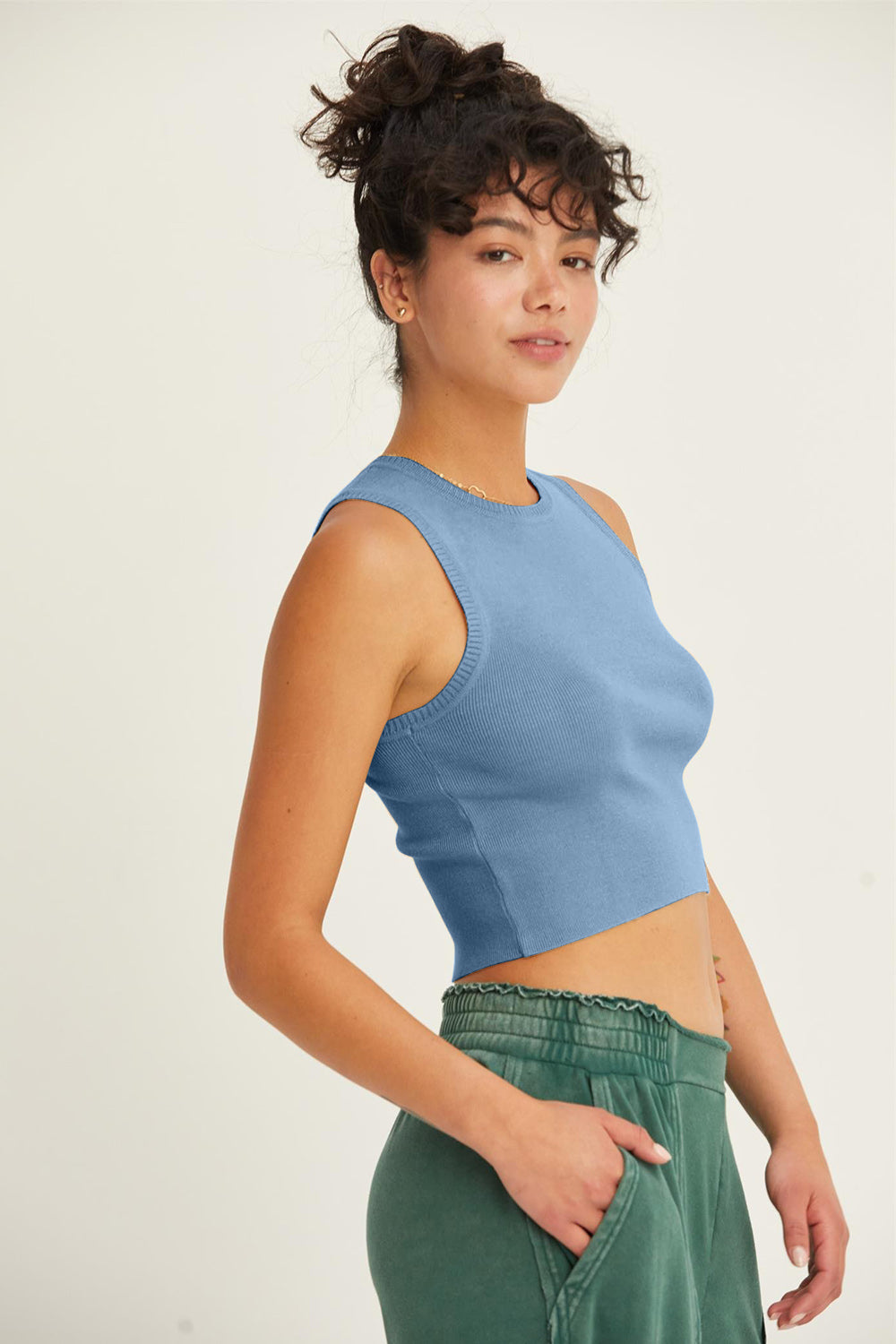 A person with curly hair is standing sideways in a casual outfit, wearing the HYFVE Ribbed Knit Cropped Tank in blue and green pants, looking toward the camera against a plain background.