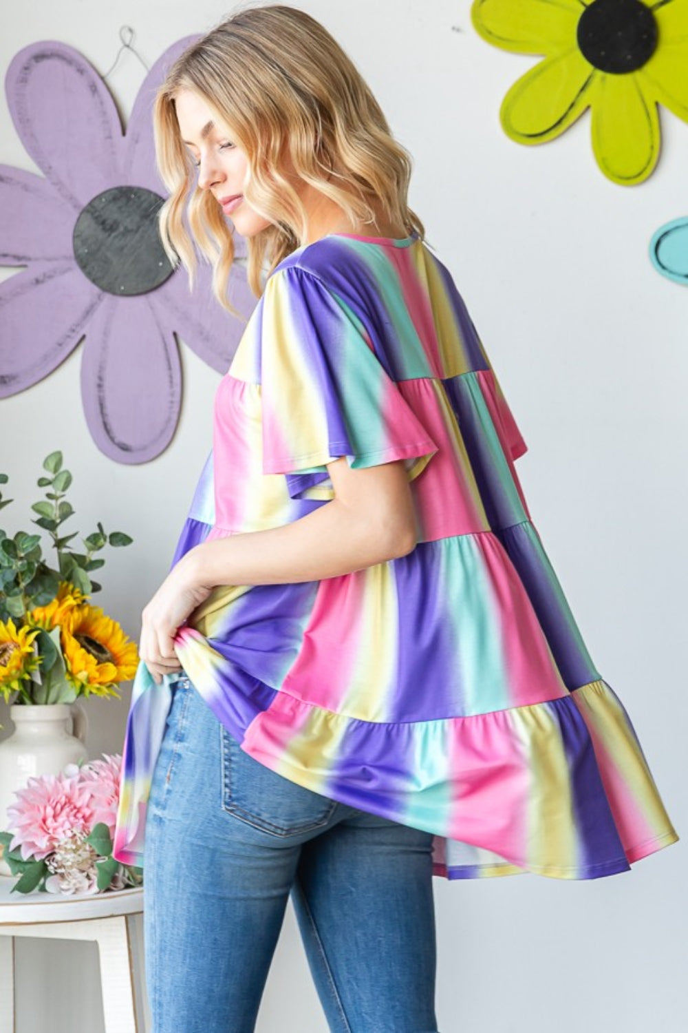 A person poses in a striking Heimish Full Size Short Sleeve Striped Tiered Top, surrounded by decorative flowers and wall accents.