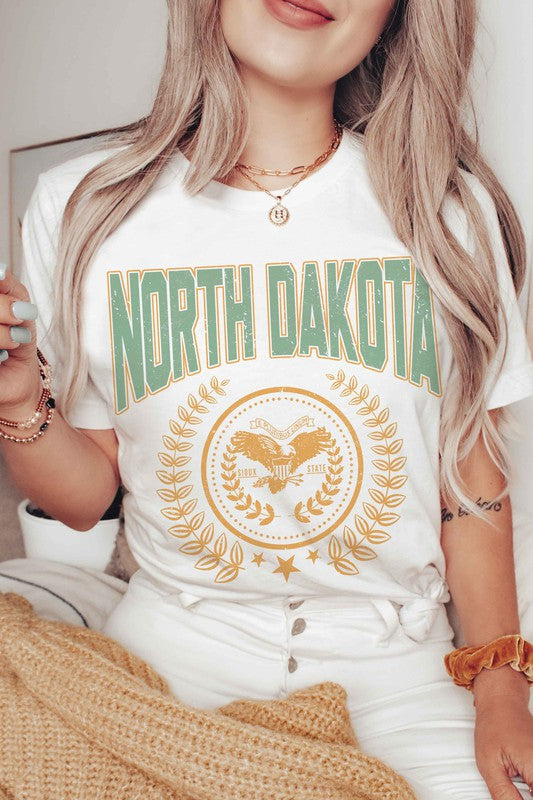 Person wearing a "NORTH DAKOTA Graphic Tee" featuring green and yellow text and graphics, holding a small object in their right hand.