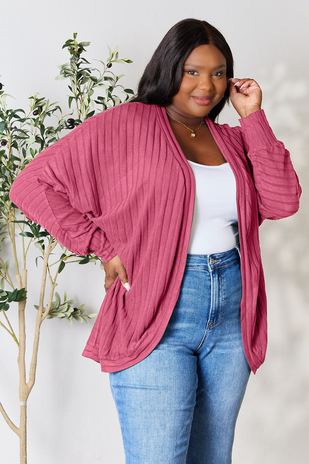 A person with wavy hair, wearing the Basic Bae Full Size Ribbed Cocoon Cardigan over a white top and beige pants, stands in front of a light-colored background with a leafy plant, showcasing an effortlessly basic style.