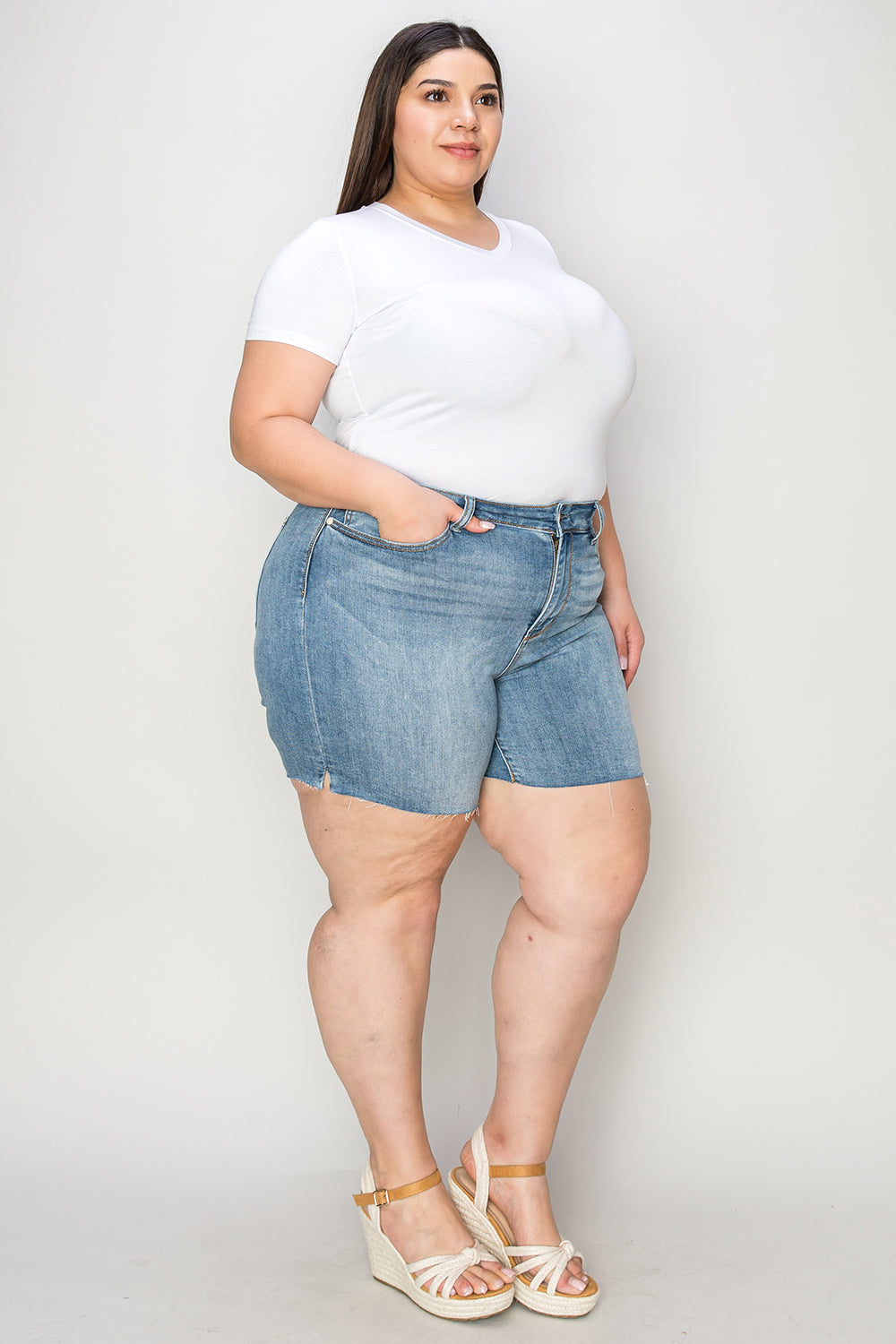 Side view of a person wearing a white shirt and Judy Blue Full Size High Waist Raw Hem Denim Shorts, standing indoors near a wooden cabinet. Summer fashion vibes radiate through the casual ensemble.