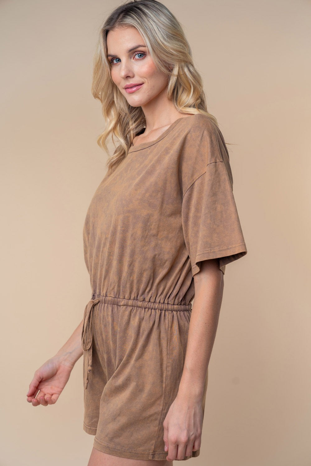 A person wearing the White Birch Full Size Short Sleeve Drawstring Romper stands against a beige background, looking slightly to the side. Perfect for your summer wardrobe, this stylish ensemble is both comfortable and chic in a cotton knit texture.