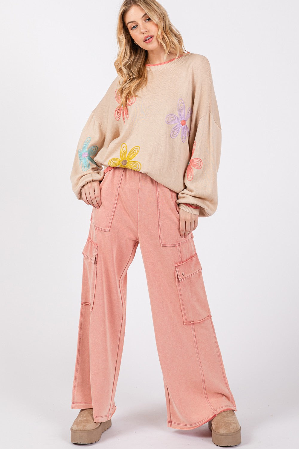 A person wearing a beige sweater adorned with colorful flower designs and pink SAGE + FIG Knit Terry Mineral Wash Wide Leg Pants stands against a plain background.