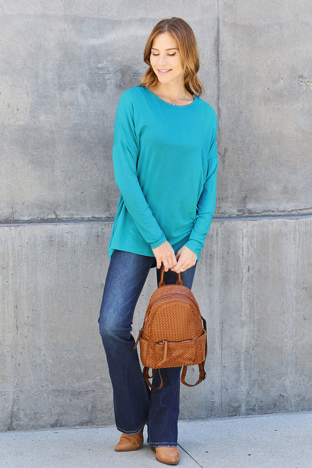 A woman with shoulder-length hair wearing a Basic Bae Full Size Round Neck Dropped Shoulder T-Shirt in green and a pair of jeans, exuding a basic style, stands against a concrete wall. She's holding an imported brown woven backpack on her shoulder while touching her hair with her hand.