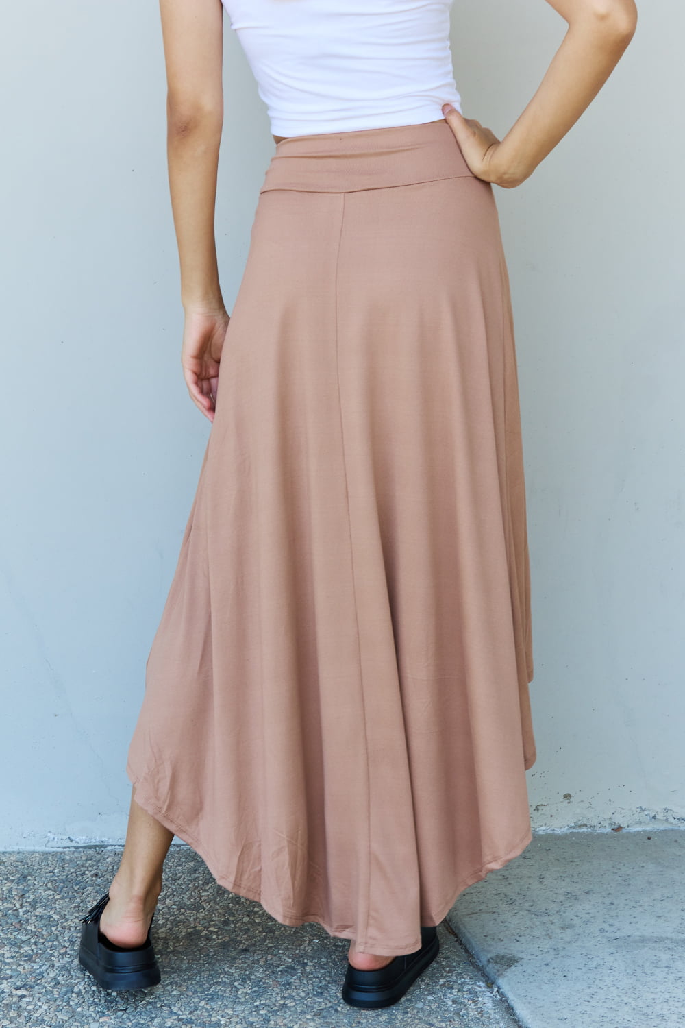 Person wearing a white crop top, the Ninexis First Choice High Waisted Flare Maxi Skirt in Camel with an asymmetrical hemline, and black sandals, standing against a light-colored wall.