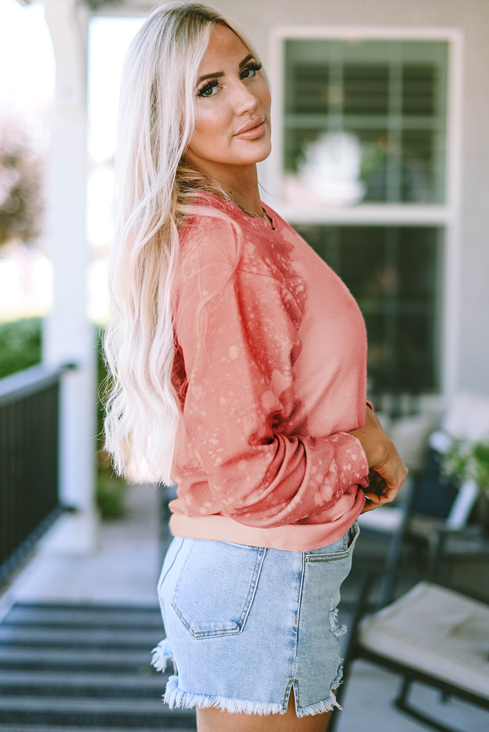 A person with long blonde hair stands with their back to the camera, wearing a Pink Bleached Round Neck Pullover Sweatshirt and light blue denim shorts—perfect for casual occasions.