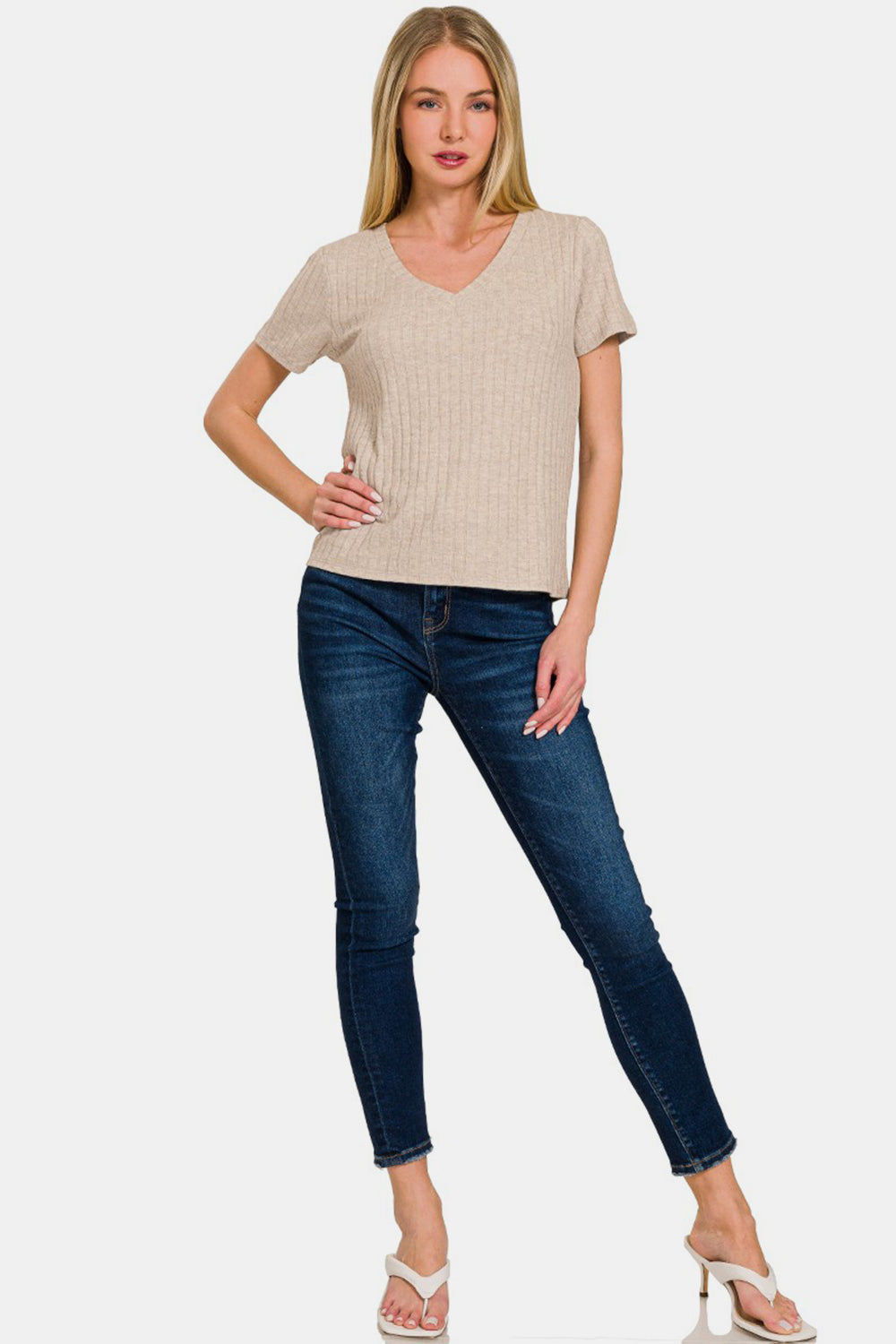 A woman with long blonde hair is wearing a high-quality beige Zenana Ribbed Short Sleeve T-Shirt and blue jeans. She has one hand on her hip.