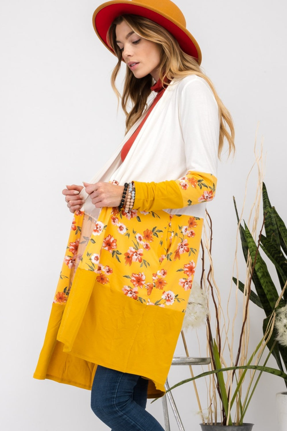 Wearing the Celeste Full Size Floral Color Block Open Front Cardigan with a white and yellow floral print, along with a red hat, a red top, and jeans, a woman stands beside a houseplant.