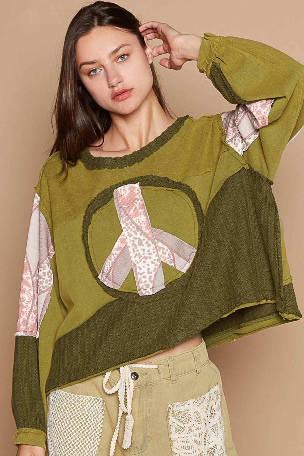 A woman with long hair is standing against a beige background, wearing the POL Long Sleeve Cut Sew Peace Emblem Top in olive green and beige pants.