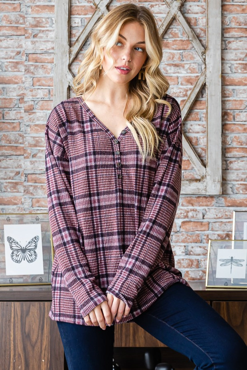 Sporting long blonde hair, a person stands against a brick wall wearing the Heimish Full Size Plaid V-Neck Long Sleeve Top in pink and black. Framed butterfly prints adorn the background, while this adaptable wardrobe item adds a touch of flair to any ensemble.