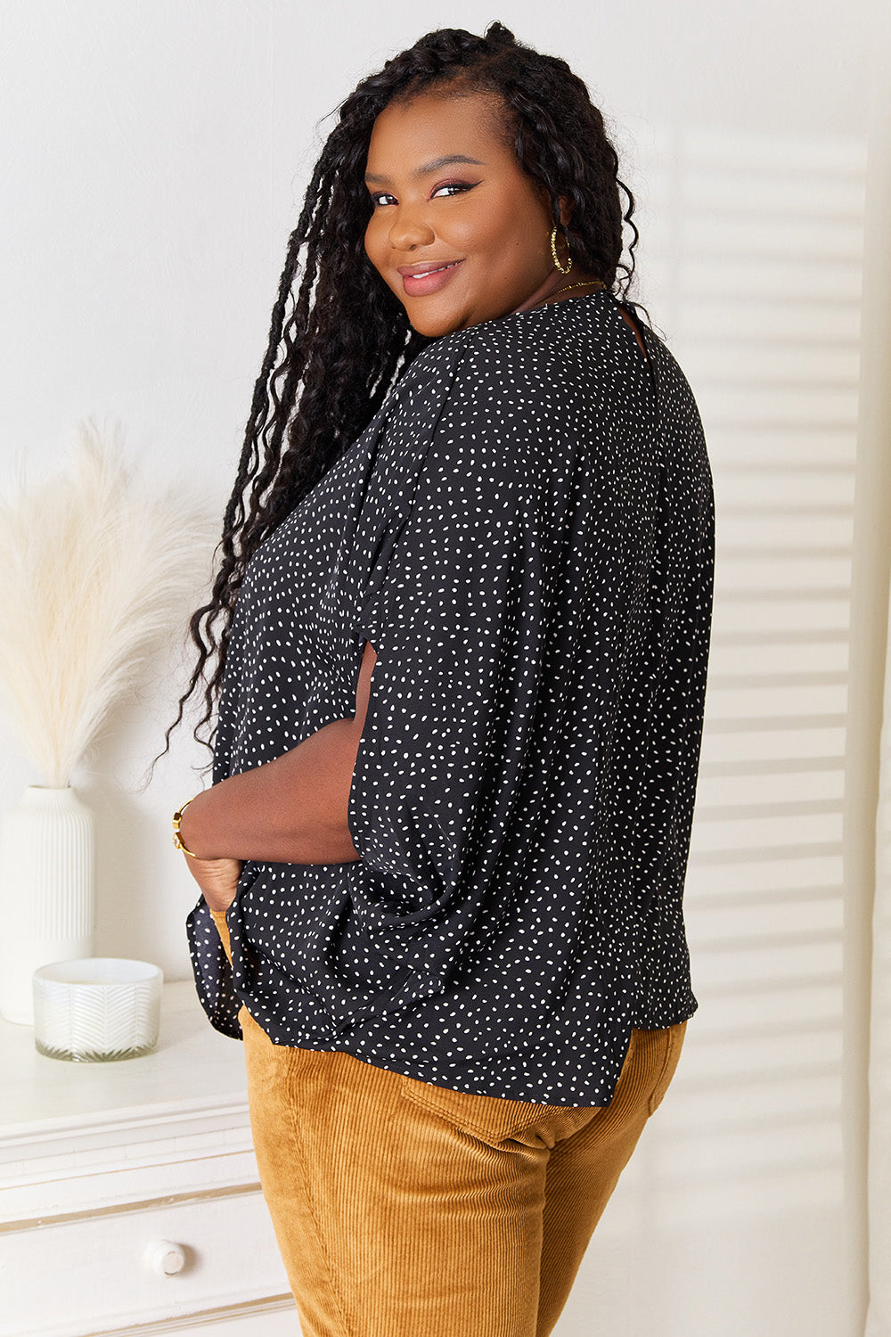 A woman stands wearing the Perfee Printed Dolman Sleeve Round Neck Blouse with white dots and black pants, smiling and looking to her left. The blouse features a printed design that adds a touch of elegance to her outfit.