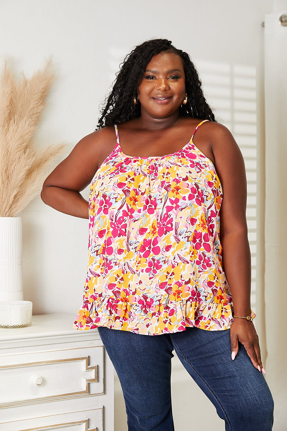 A person stands indoors, smiling with one hand on their hip, while wearing the Double Take Floral Scoop Neck Ruffle Hem Cami and blue jeans.