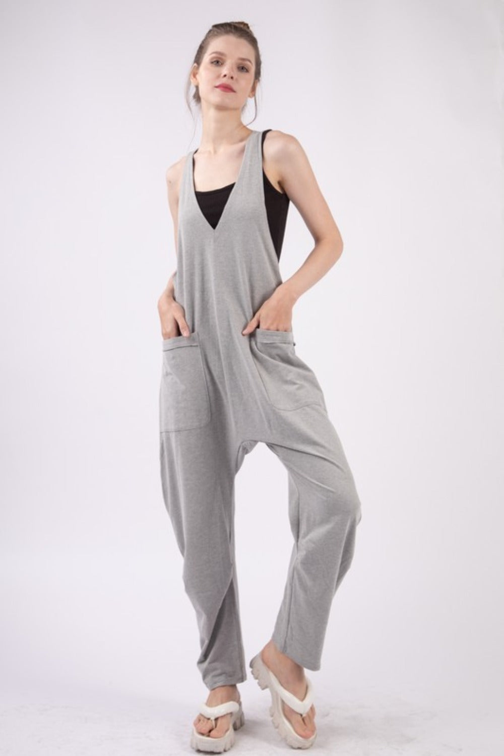 A person wearing the VERY J Plunge Sleeveless Jumpsuit with Pockets in gray and white sandals stands against a plain white background.