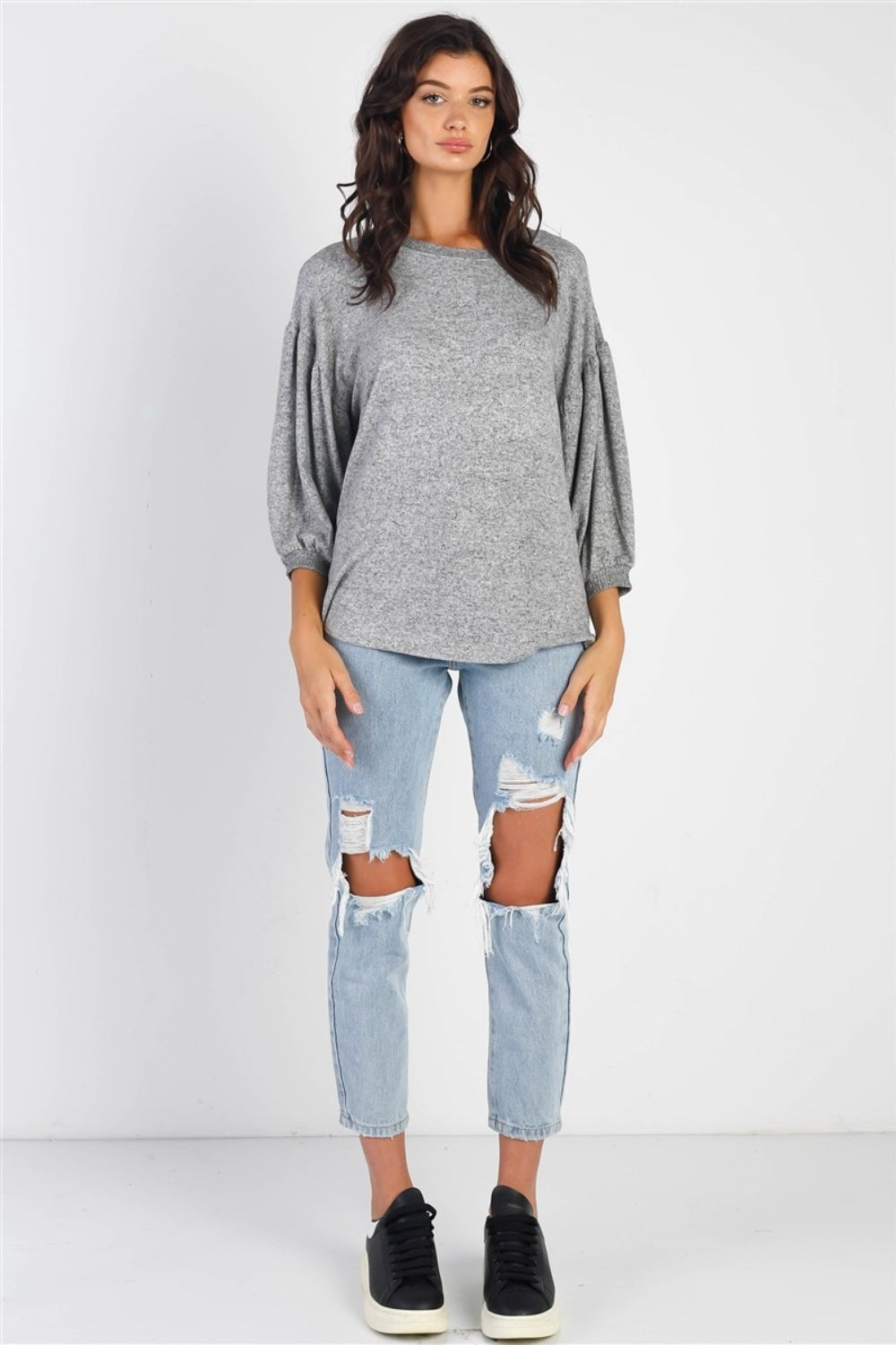 A person with long dark hair stands against a white background, smiling and raising one hand while wearing the trendy and stylish Cherish Apparel Drop Shoulder Puff Sleeve Top in burgundy paired with light blue jeans.