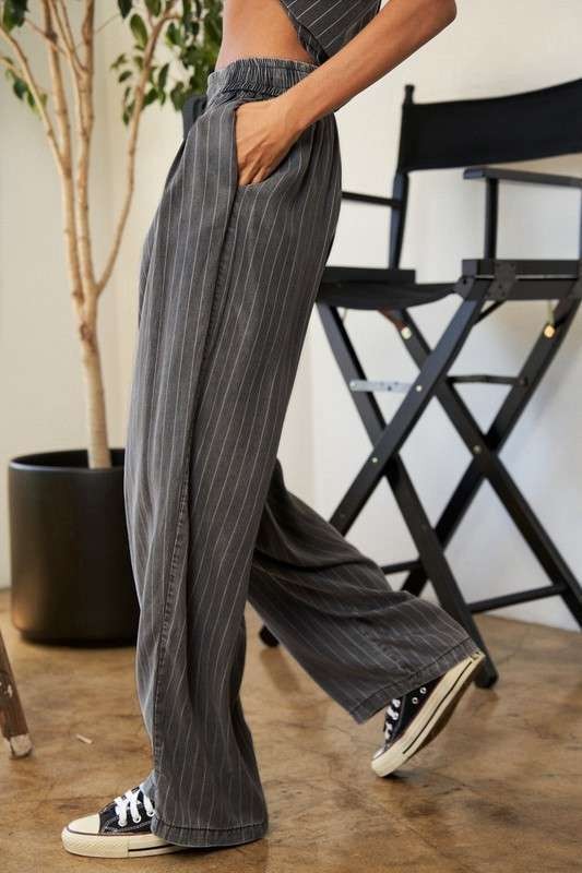 A person wears a casual outfit called the Stripe Button Down Shirt and Long Pants Set, which features a loose-fitting pinstriped design, paired with sneakers. They stand indoors near a potted plant and a director's chair.