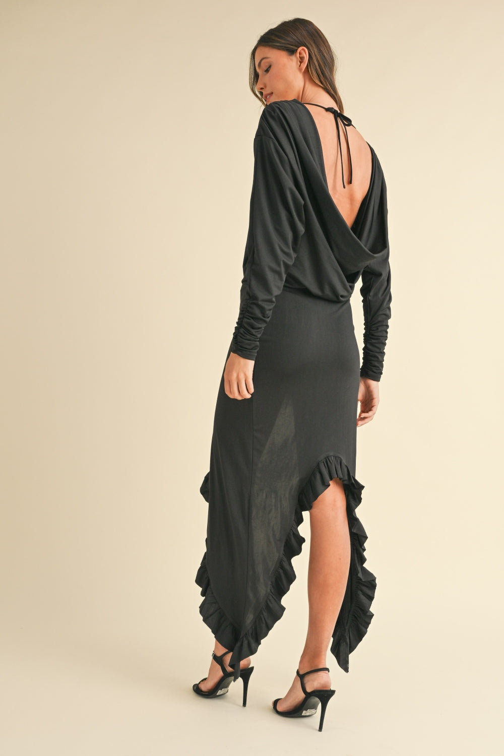 A woman is wearing the Mable Backless Asymmetric Ruffle Hem Dress, featuring a stunning black, backless design with a low back and asymmetrical hemline. She stands sideways and looks down, showcasing the long sleeves and elegant ruffle details of the dress, which she stylishly pairs with black heels.