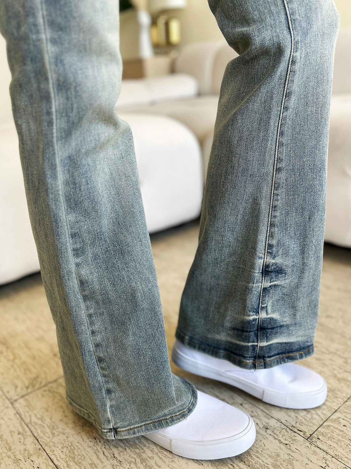 A person wearing Judy Blue Full Size Mid Rise Flare Jeans in a retro-inspired light blue color, paired with a white top and white shoes, stands in a modern living room with a white tufted sofa in the background.