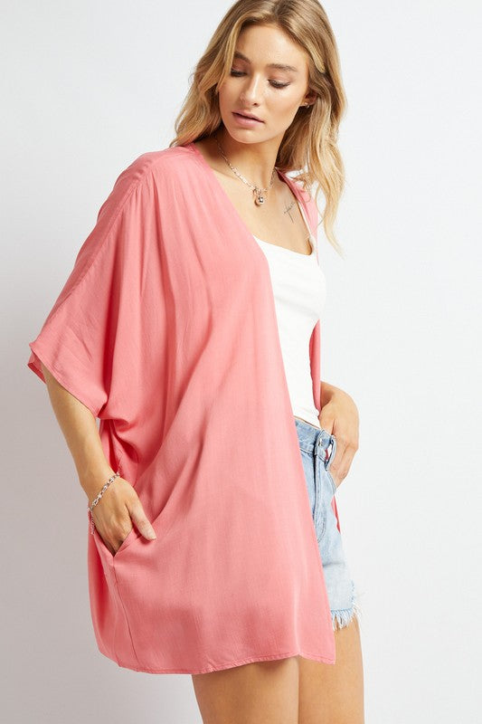 A woman poses against a simple backdrop, gracefully wearing a wardrobe essential: the Favorite Solid Kimono Cardigan in pink. Complementing her white tank top and denim shorts, this casual layer enhances her relaxed style with an elegant touch.