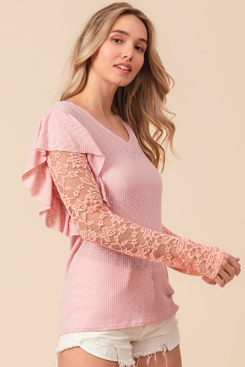 A woman with long blonde hair, smiling and looking to the side, is wearing a pink BiBi Ruffled Lace Sleeve Rib Knit Top paired with white shorts.