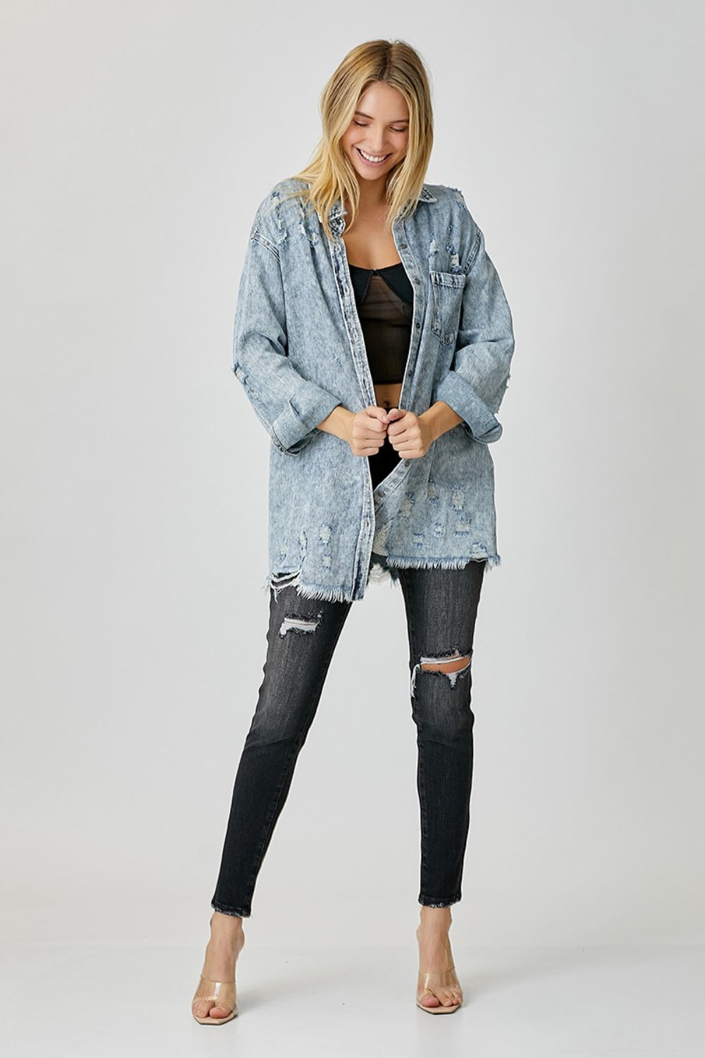 A smiling person wearing a RISEN Distressed Raw Hem Denim Shirt and black ripped jeans stands against a plain background, exuding a casual and edgy vibe.