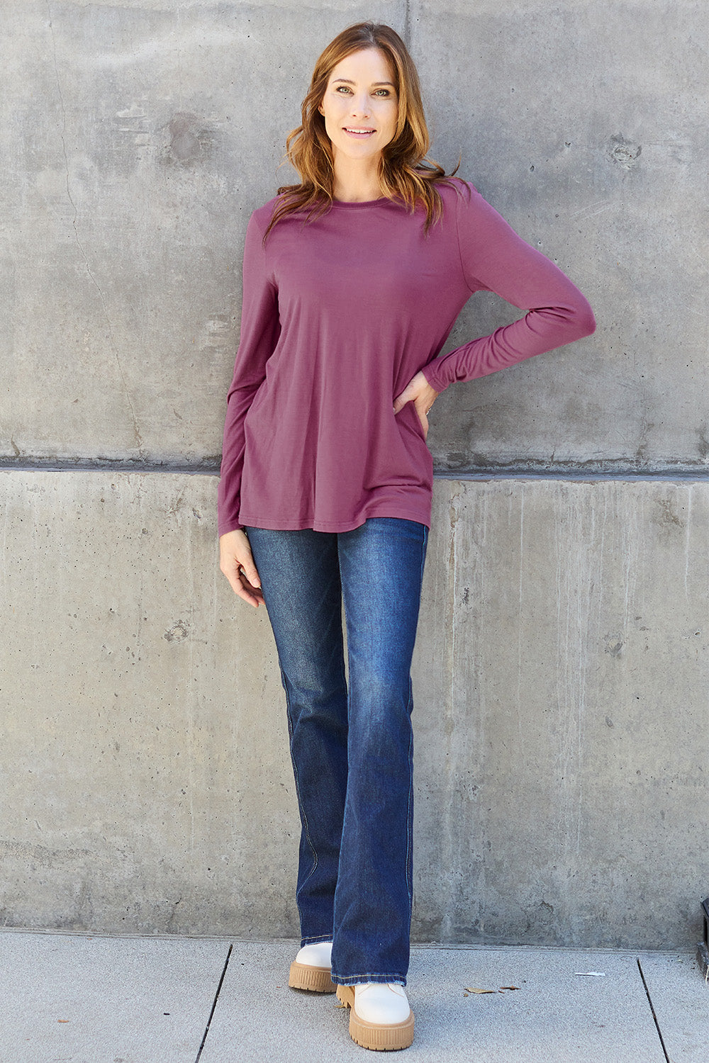 A woman with shoulder-length hair wearing the Basic Bae Full Size Round Neck Long Sleeve Top and blue jeans stands in front of a concrete wall, holding a beige handbag over her shoulder. Her outfit exemplifies basic style, with the top made from 100% polyester and recommended for machine wash cold.