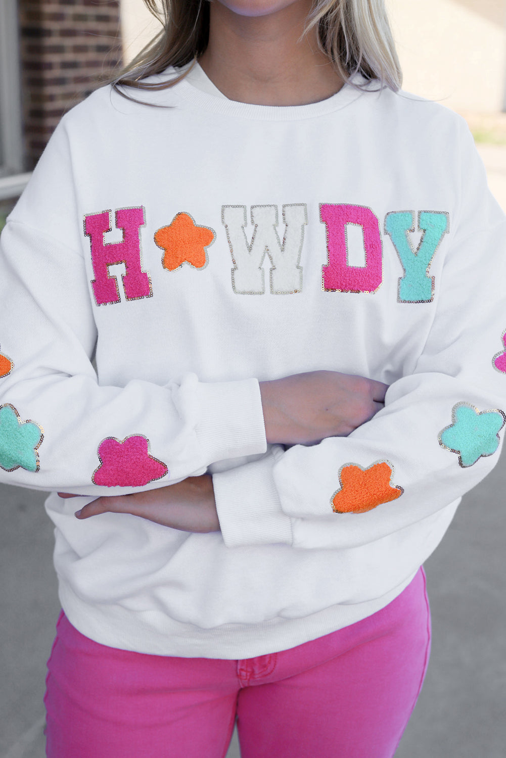 A person poses in the White Howdy Glitter Chenille Patch Graphic Casual Sweatshirt, adorned with colorful star patterns and the letters "WD." She wears light denim shorts and has shoulder-length blonde hair.