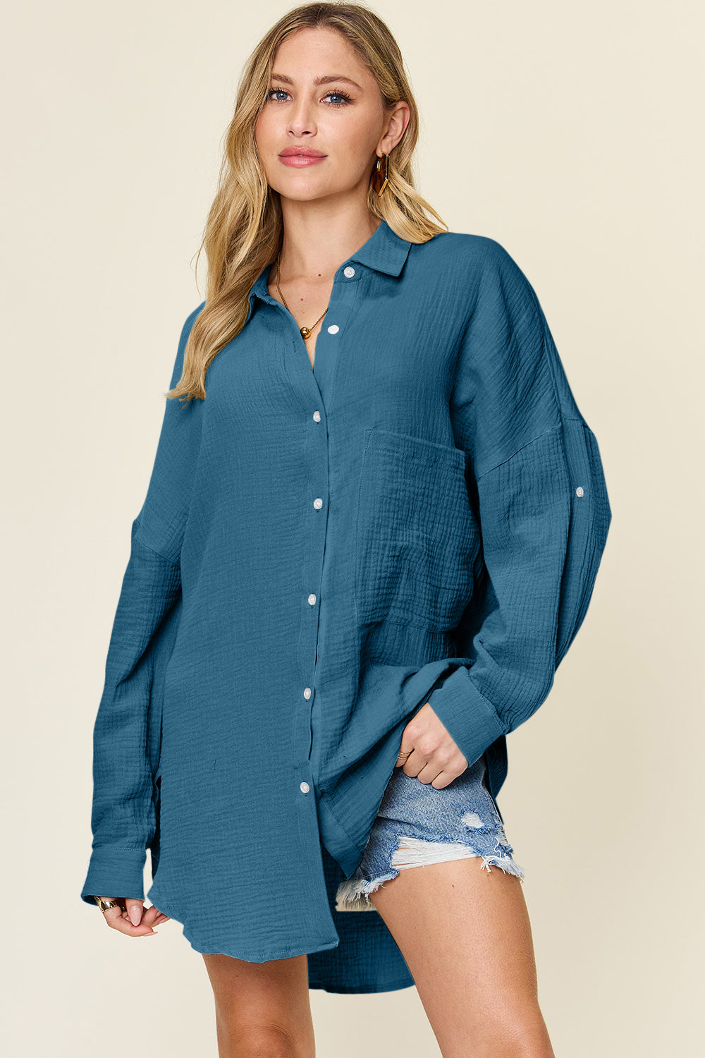 A person is wearing the Double Take Full Size Pocketed Texture Button Up Shirt, a long-sleeved, color-blocked piece made of 100% cotton in shades of green and cream. They have paired it with distressed denim shorts and are standing against a neutral backdrop. The shirt features buttoned cuffs and offers a relaxed yet stylish look. It is easy to care for with machine wash cold instructions.