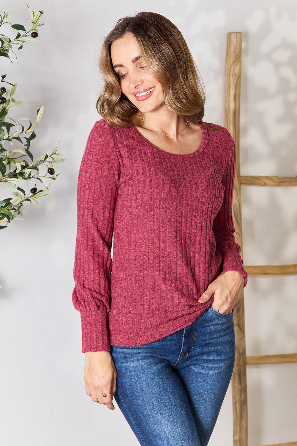 A person in a stylish maroon Ribbed Round Neck Lantern Sleeve Blouse and jeans stands by a wooden ladder, with olive branches in the background.