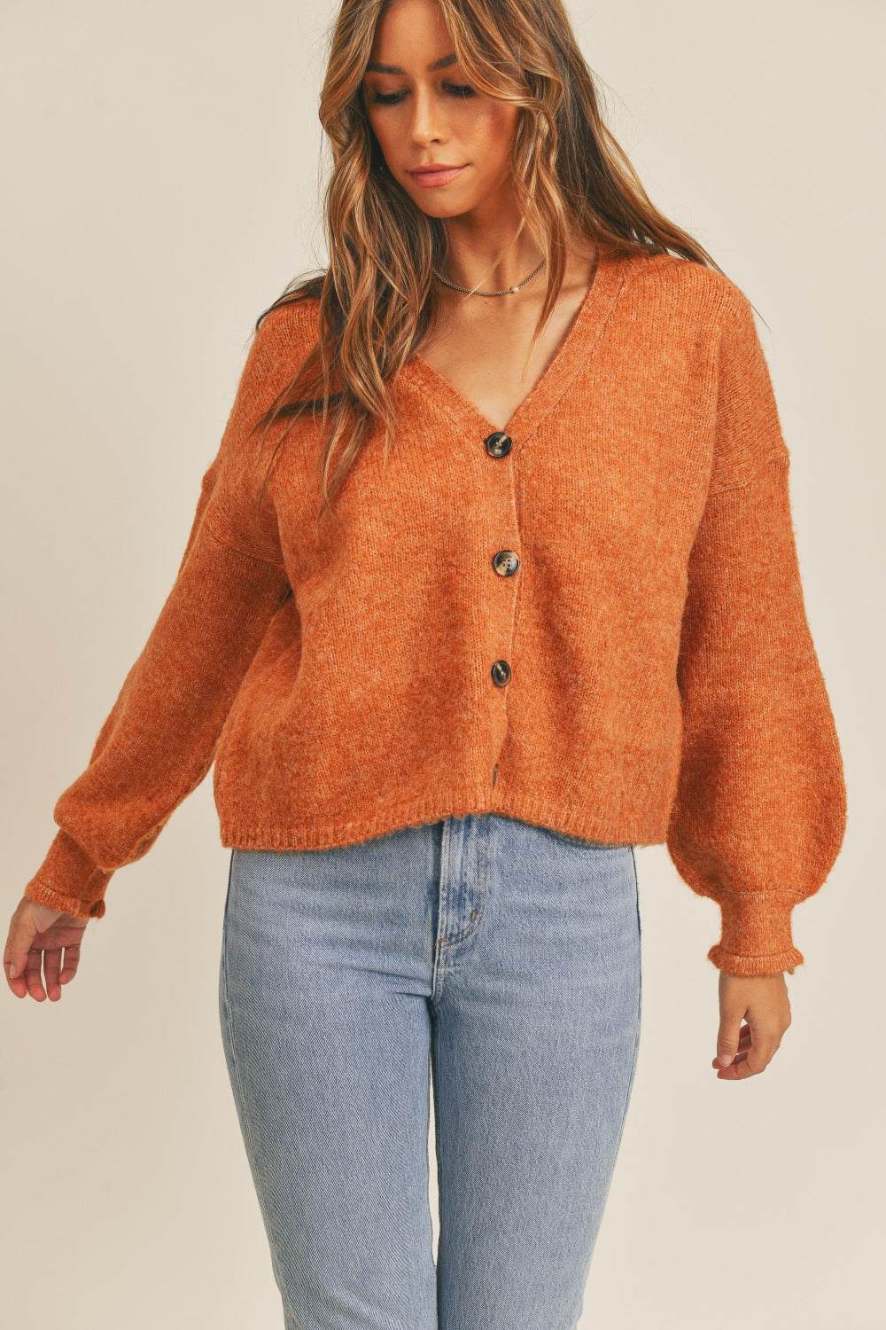 A woman wearing a MABLE Long Sleeve Button Down Sweater Cardigan in burnt orange and light blue jeans looks to her left against a plain background, exuding classic elegance.