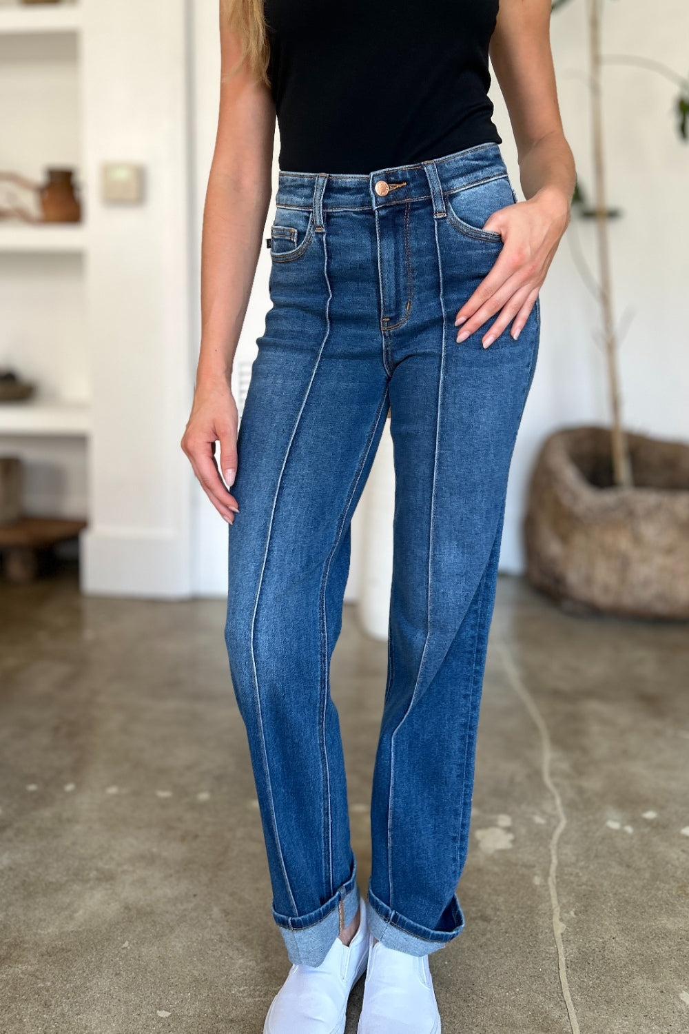 Two women stand side by side indoors, both wearing black tank tops, Judy Blue Full Size High Waist Front Seam Detail Straight Jeans, and white shoes. They are smiling and posing for the camera. A plant and shelves with decorations are in the background.