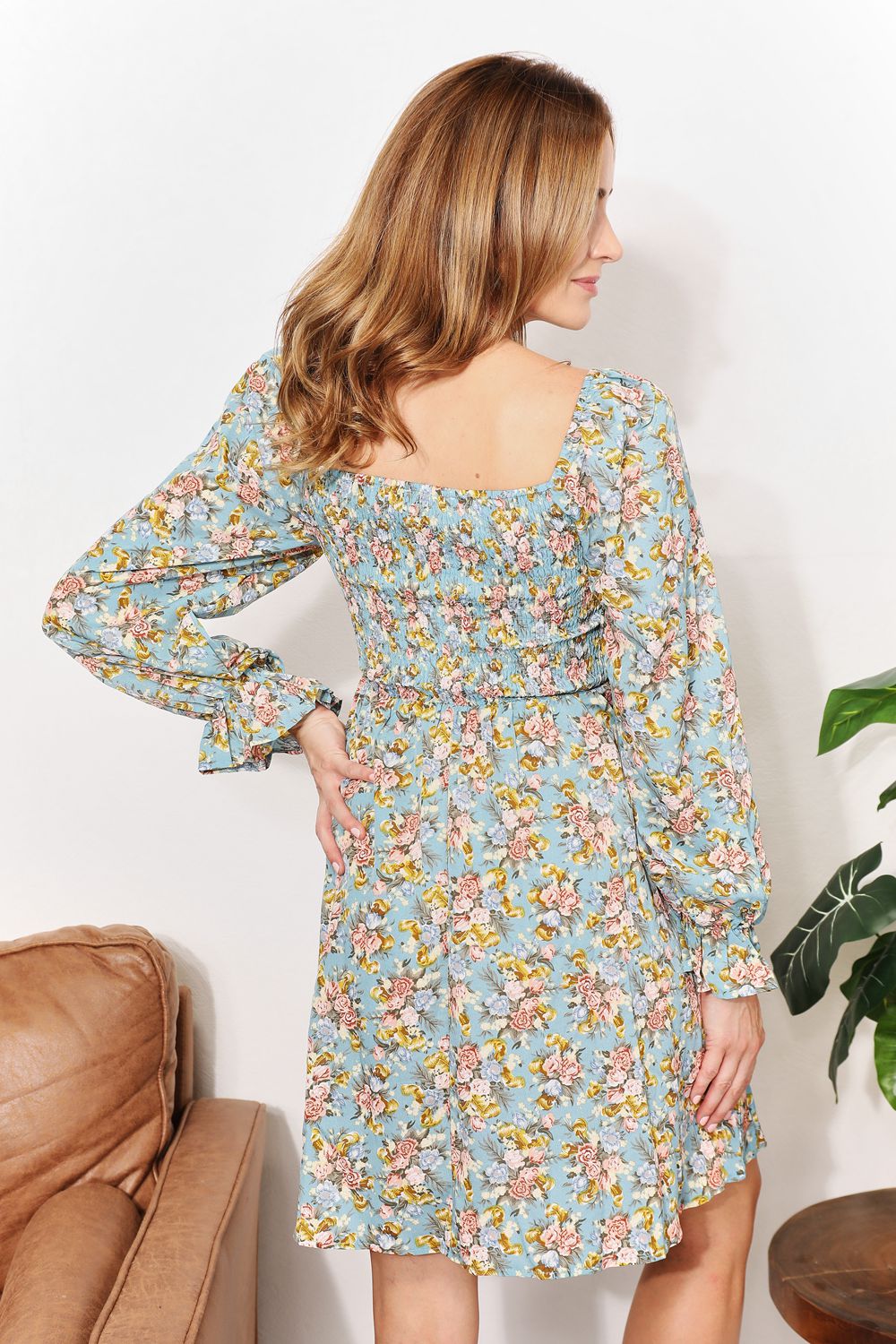 A woman in the Honey Floral Smocked Flounce Sleeve Square Neck Dress stands indoors, holding the hem of her dress. She is smiling slightly with her eyes looking downward. Nearby, a plant and a brown couch complete the cozy scene.