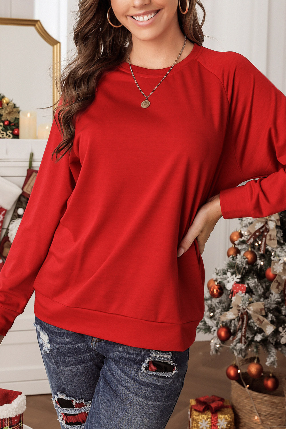 A person wearing a cozy Fiery Red Solid Round Neck Raglan Sleeve Sweatshirt and jeans stands indoors with a decorated Christmas tree and wrapped presents in the background.