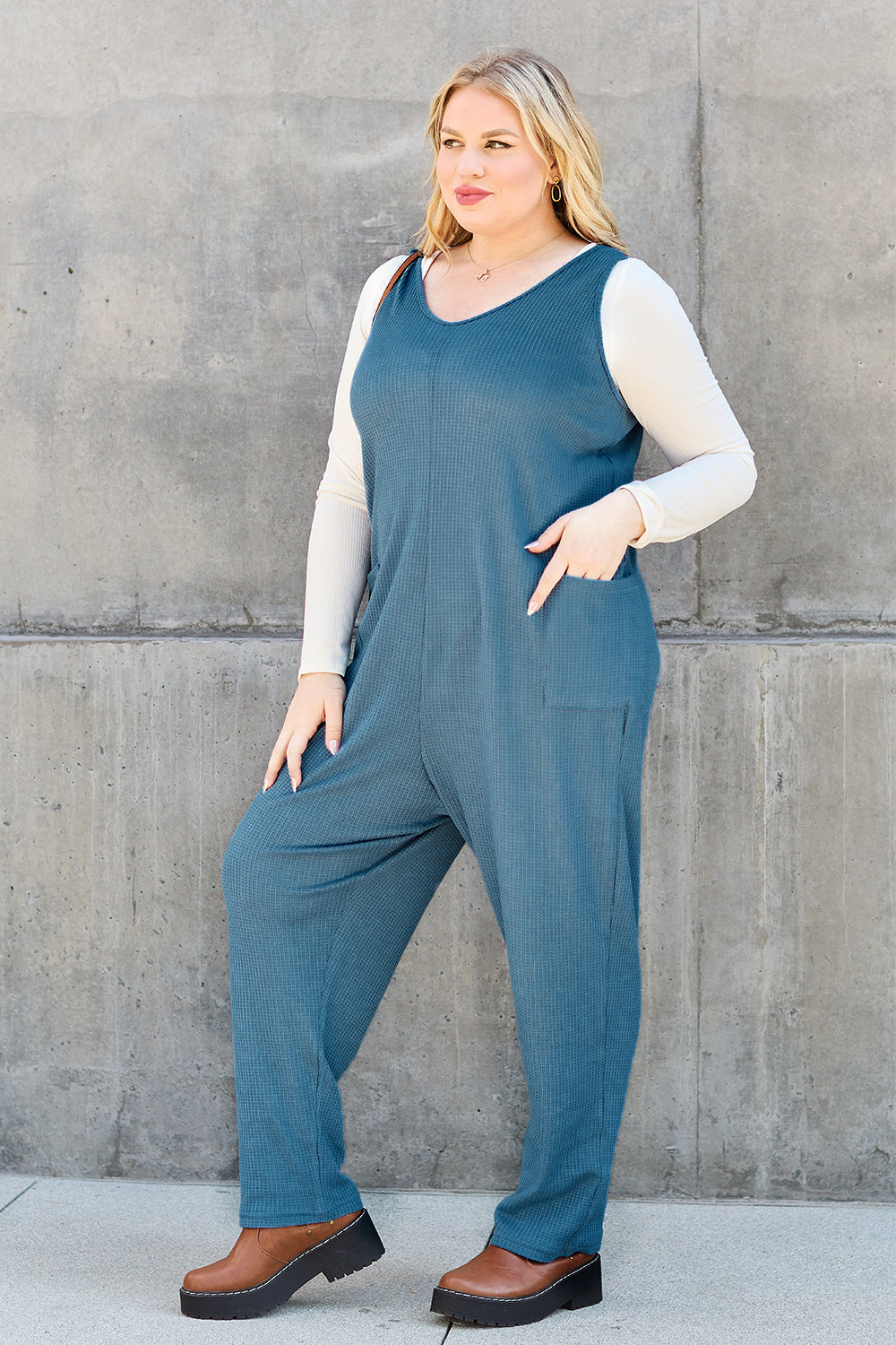 A person stands outdoors wearing the Double Take Full Size Sleeveless Straight Jumpsuit in blue and brown sandals, holding a cream-colored bag. In the background, there's a wooden bench and palm trees.