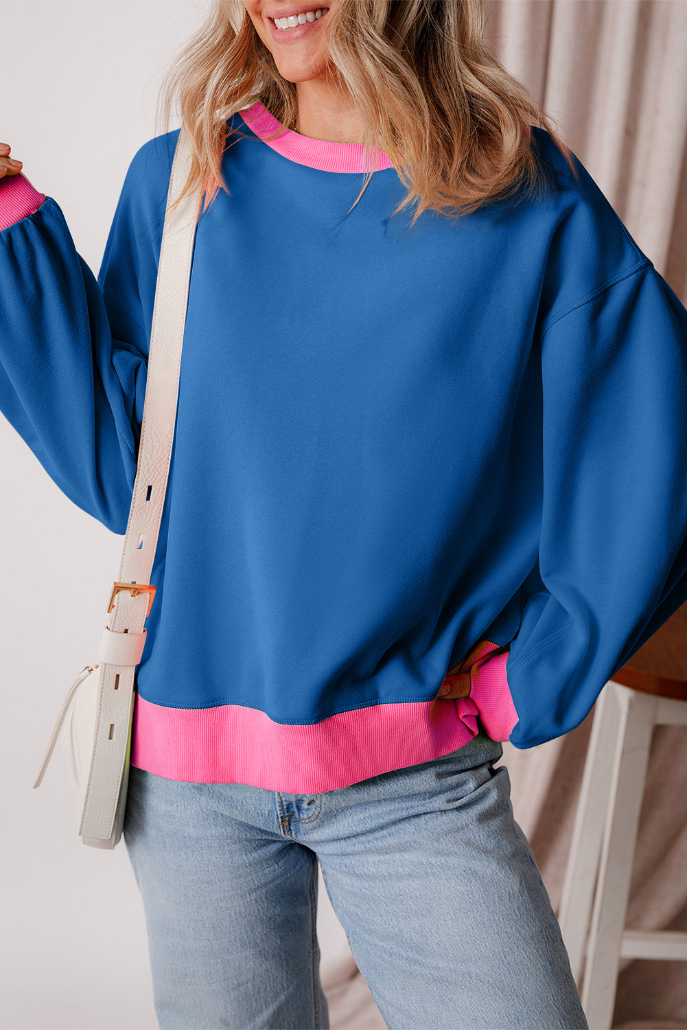 A woman with long blonde hair is smiling as she poses against a purple background, wearing the Blue Colorblock Bubble Sleeve Sweatshirt paired with white pants. The sweatshirt features a boxy fit with pink accents and is ideal for the autumn and winter seasons.