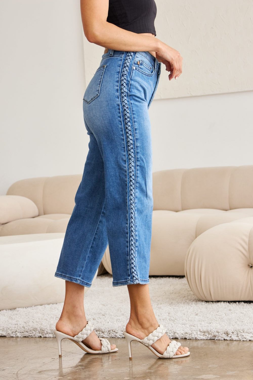 A person wearing Judy Blue Full Size Braid Side Detail Wide Leg Jeans and black heels is standing on a polished floor. There is a white couch and a large potted plant in the background.