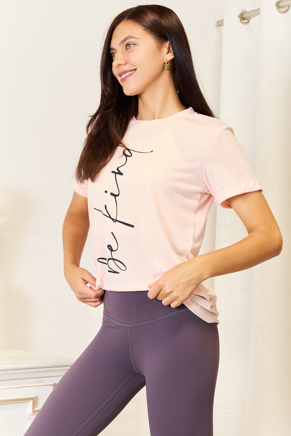 A woman wearing a light pink Simply Love BE KIND Graphic Round Neck T-Shirt with the phrase "be kind" written in cursive. She is standing indoors with a neutral expression, the round neck adding a touch of casual elegance.