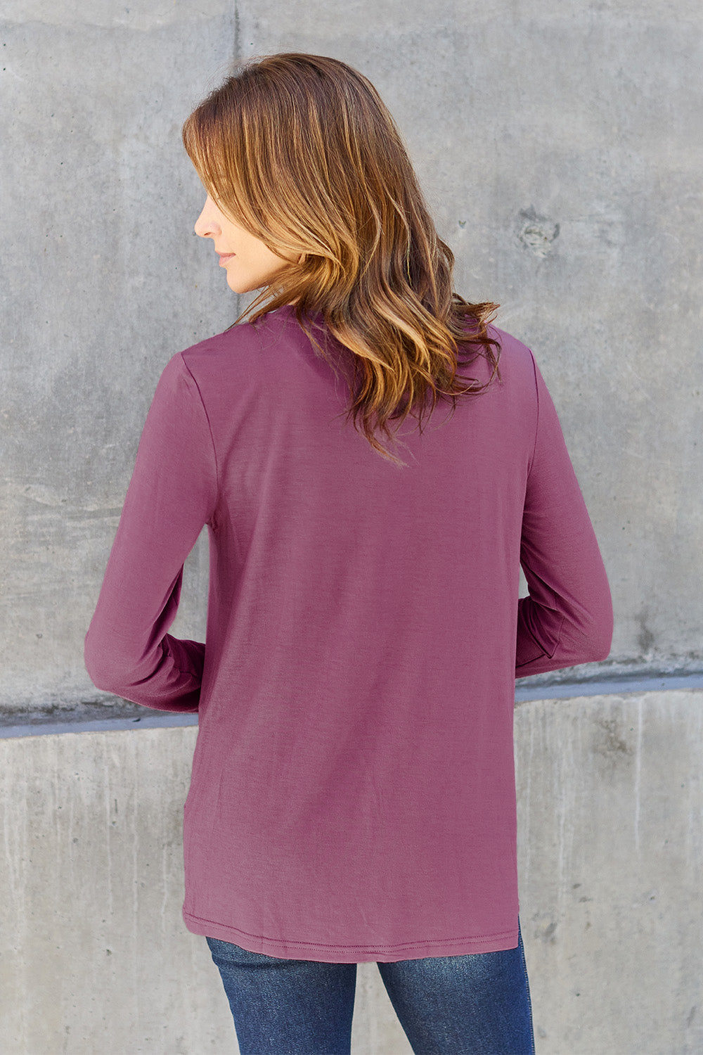 A woman with shoulder-length hair wearing the Basic Bae Full Size Round Neck Long Sleeve Top and blue jeans stands in front of a concrete wall, holding a beige handbag over her shoulder. Her outfit exemplifies basic style, with the top made from 100% polyester and recommended for machine wash cold.