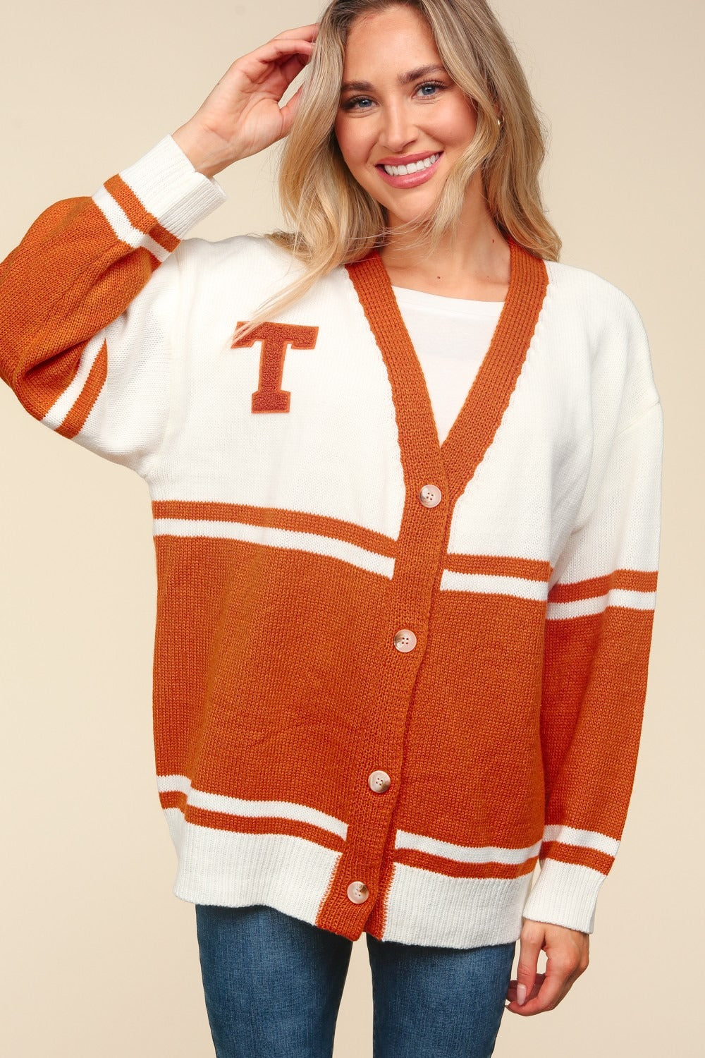 A smiling woman wearing the Haptics Full Size V Neck Button Down Letter Patch Cardigan, an oversized fit in white and orange with a "T" patch, poses with one hand on her hip and the other touching her hair.