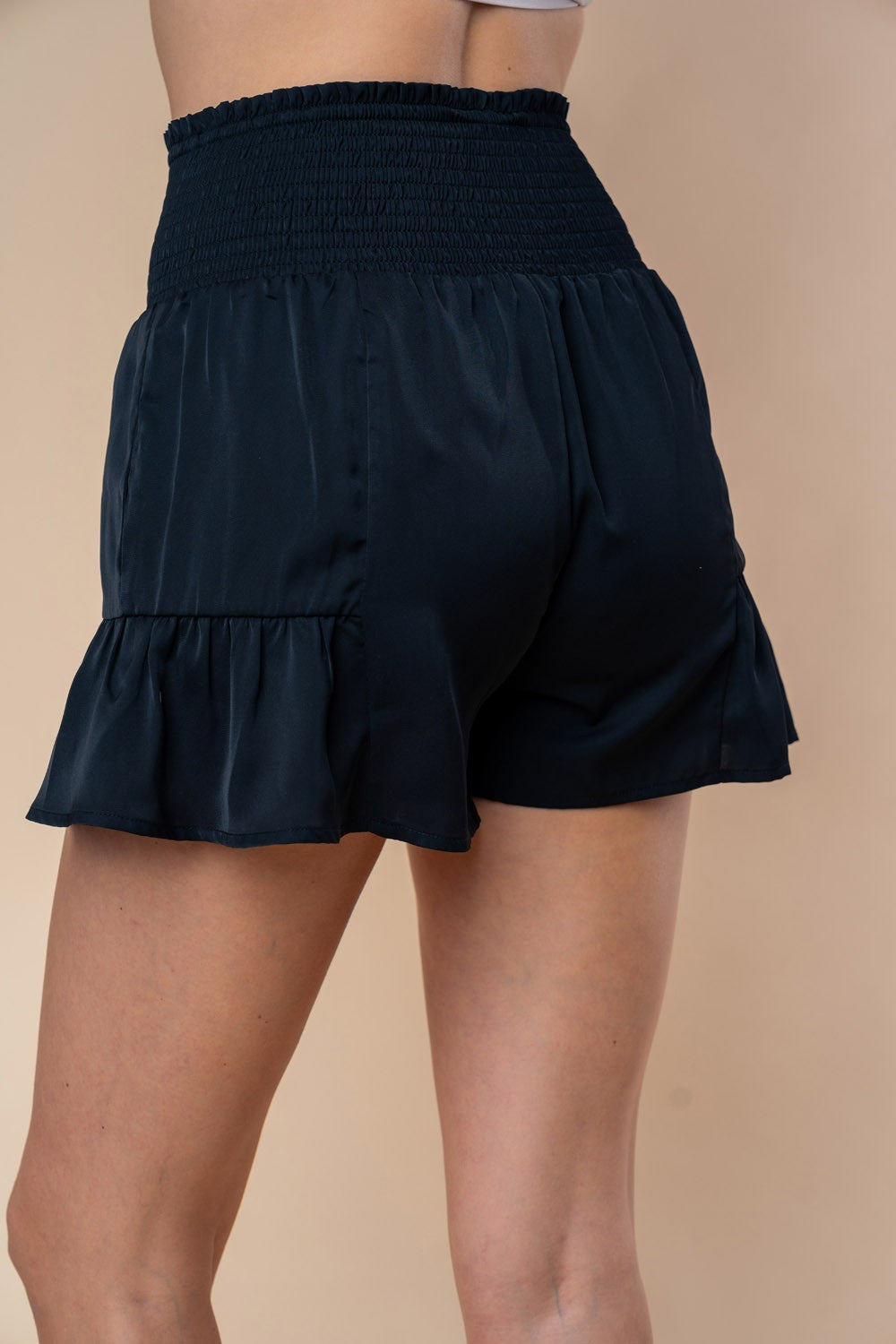 A woman is wearing the White Birch Full Size High Waisted Smocked Shorts, which feature a smocked elastic waistband with a large front bow and charming ruffle hem detailing.