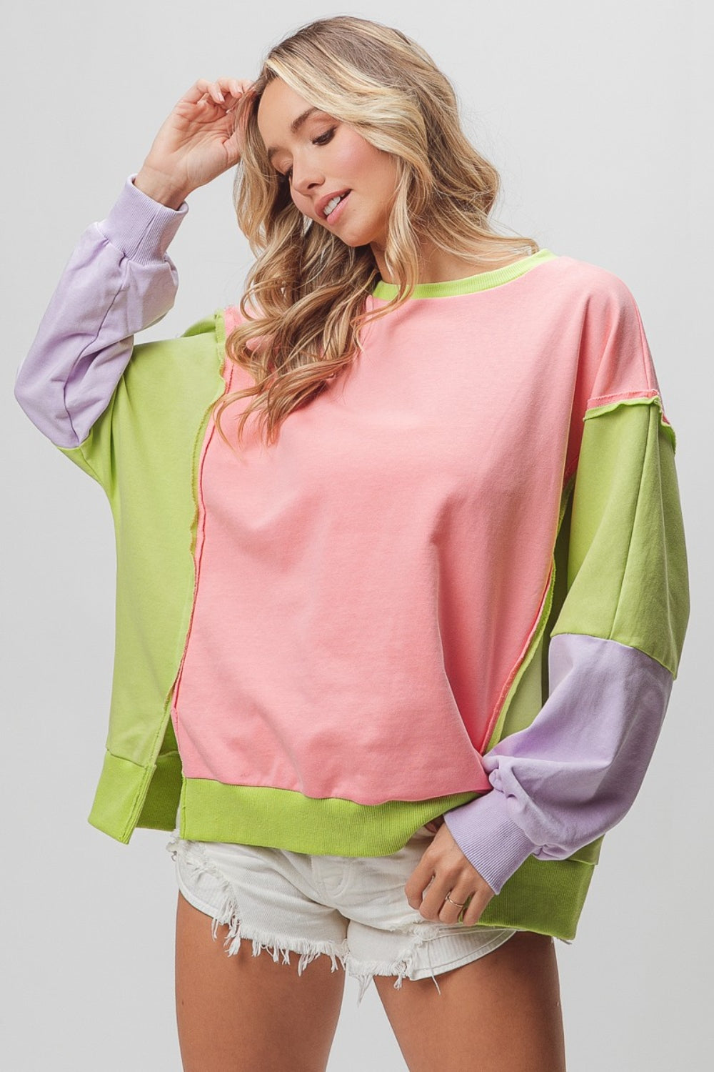 A woman is wearing a BiBi Washed Color Block Sweatshirt featuring vintage-inspired sections in pink, green, and purple. She has paired it with white distressed shorts and is posing with one arm slightly raised, perfectly showcasing the relaxed fit.