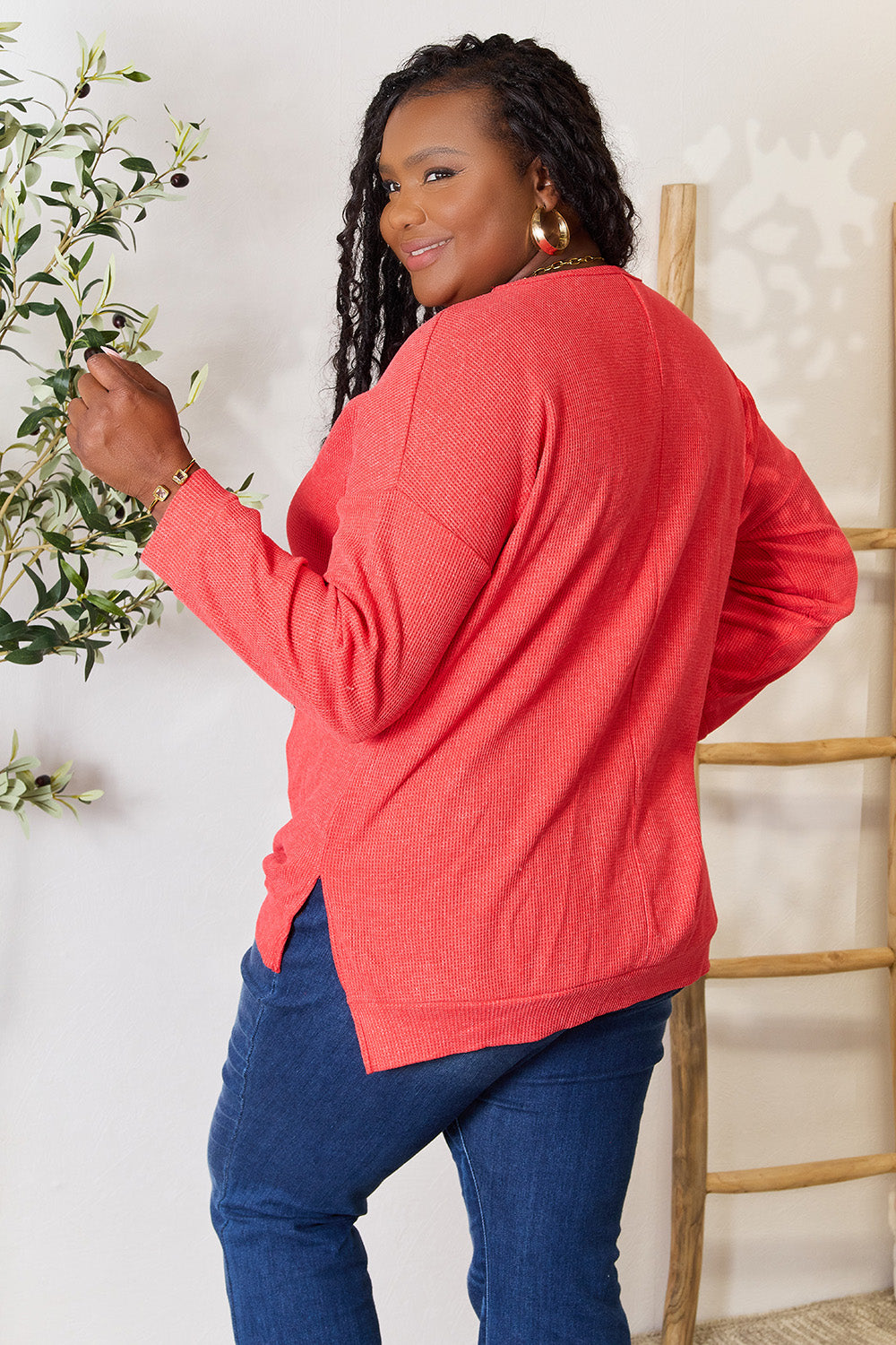 A woman wearing the Basic Bae Round Neck Drop Shoulder Slit Sweatshirt in red and blue jeans stands indoors, smiling and touching her hair, with a decorative plant and wooden ladder in the background.