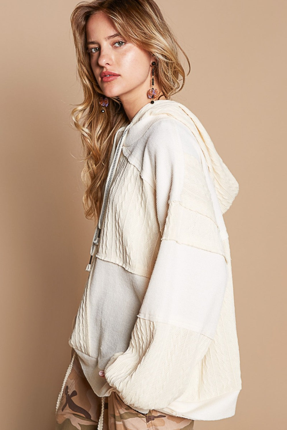 A person wearing a POL Exposed Seam Hooded Knit Top and camouflage pants sits on a stool against a beige background.