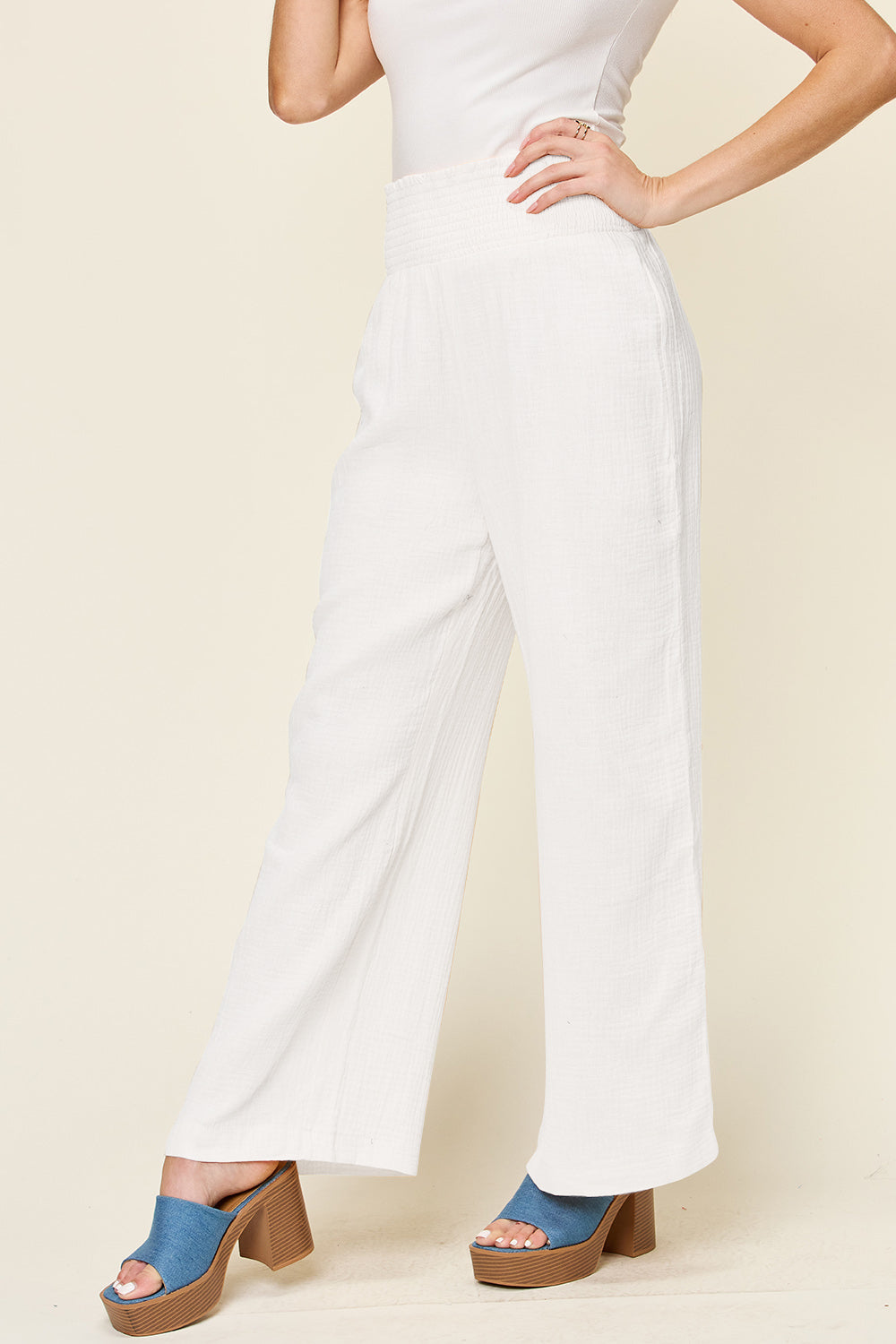 A person wearing Double Take Full Size Texture Smocked Waist Wide Leg Pants in black and a white 100% cotton top is standing with one hand on their hip. They are also wearing blue open-toe sandals.