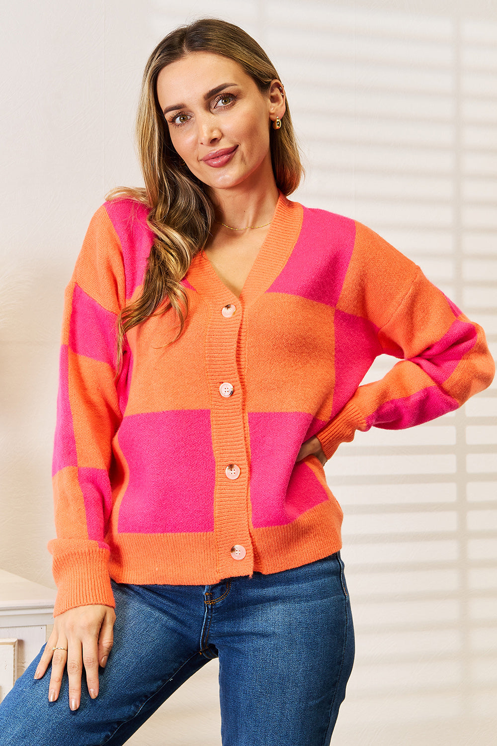 A woman with long hair stands smiling, wearing the Angel Wings Woven Right Checkered V-Neck Dropped Shoulder Cardigan in pink and orange over blue jeans.