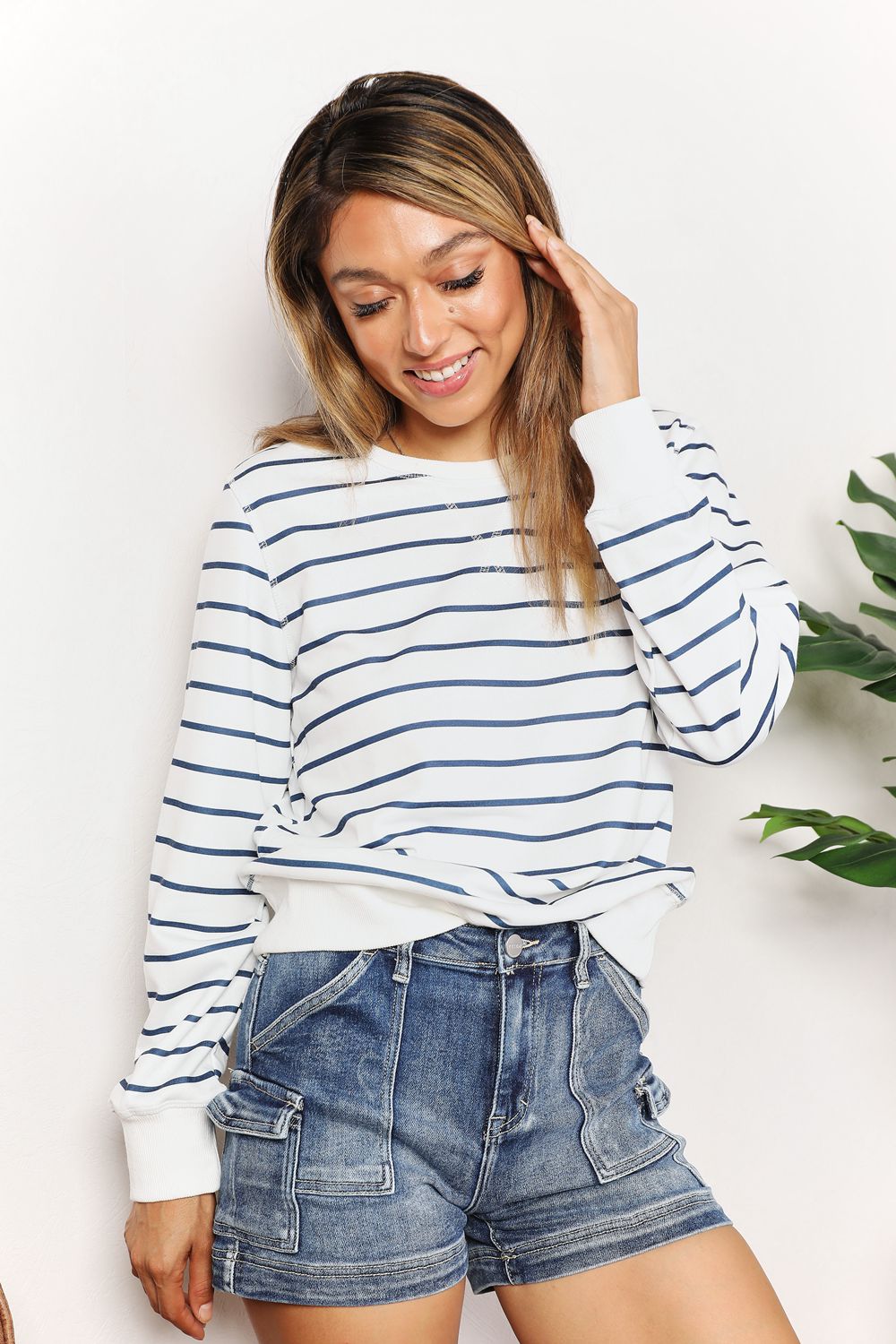 A person smiling while wearing the Double Take Striped Long Sleeve Round Neck Top, paired with denim shorts, stands in front of a white background with a plant.