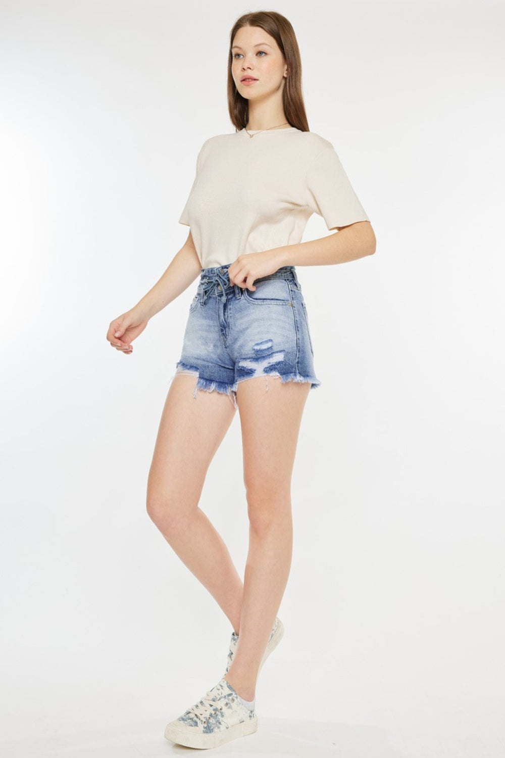 A person wearing a beige t-shirt and Kancan High Rise Frayed Hem Denim Shorts, paired with patterned sneakers, stands against a white background—an ideal addition to any summer wardrobe.