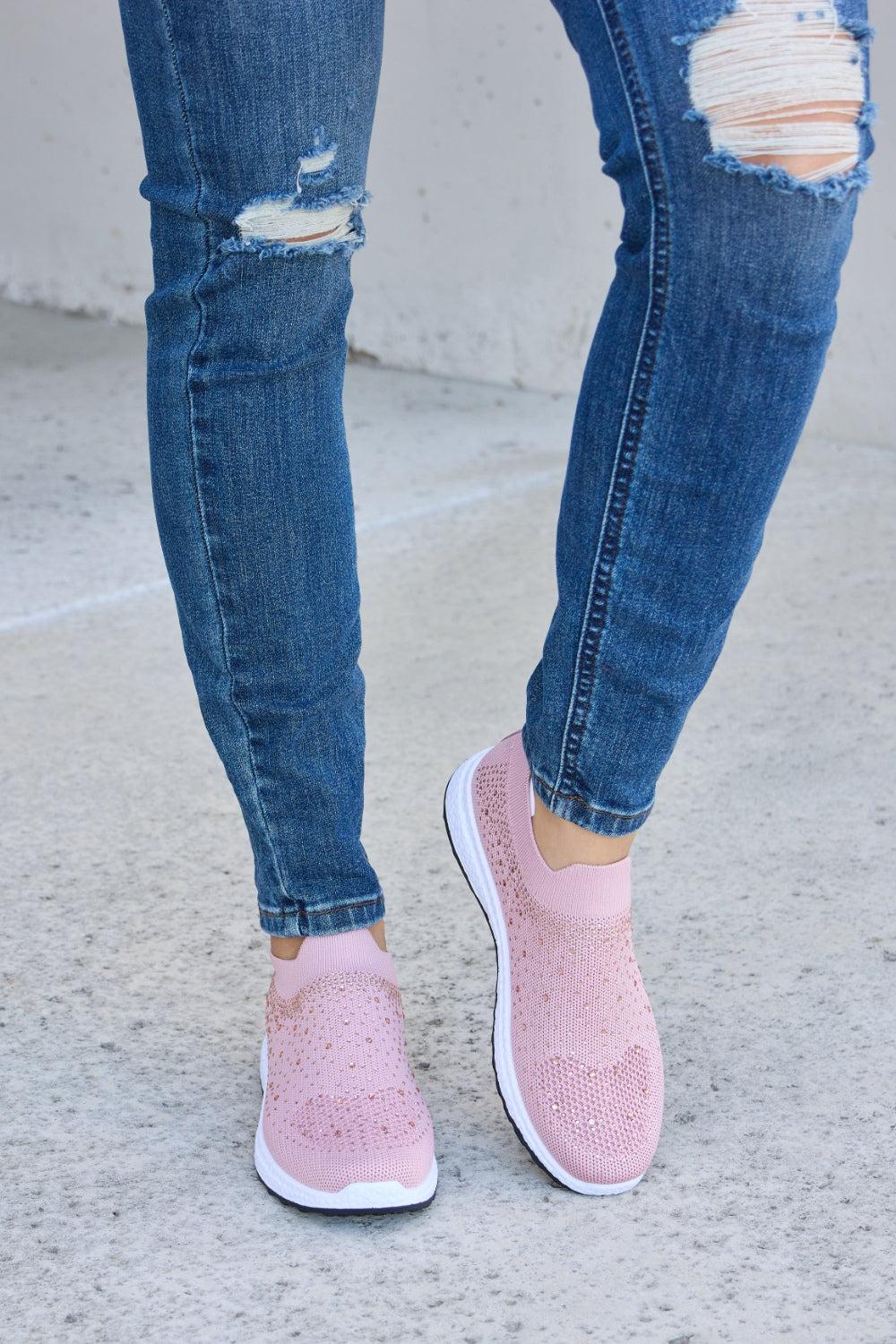 Person wearing Forever Link Rhinestone Mesh Breathable Sneakers in light pink and ripped blue jeans, standing on a concrete surface.