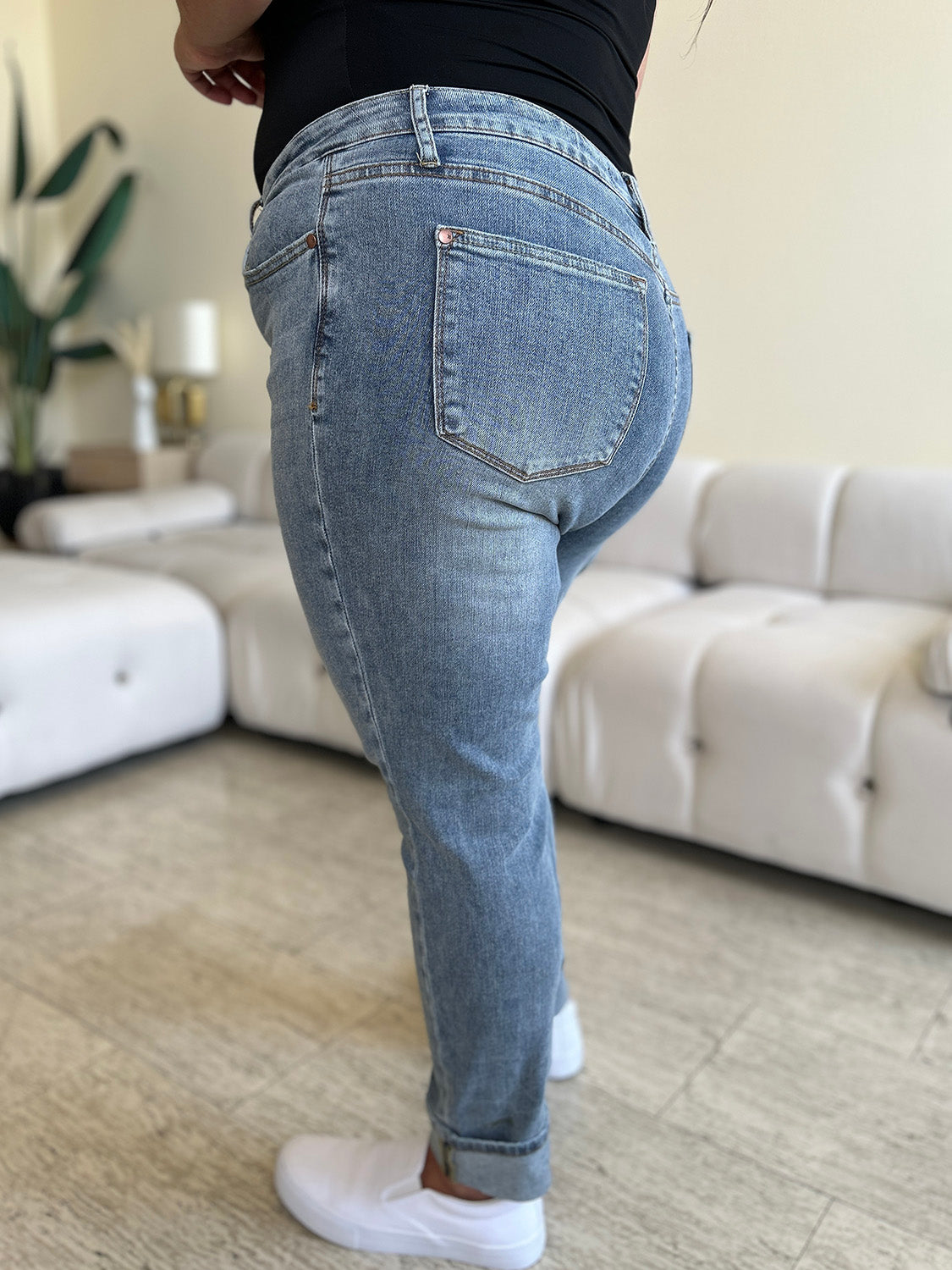 A person wearing Judy Blue Full Size High Waist Cuff Hem Jeans and white slip-on shoes stands in a living room with a white couch and plants in the background.