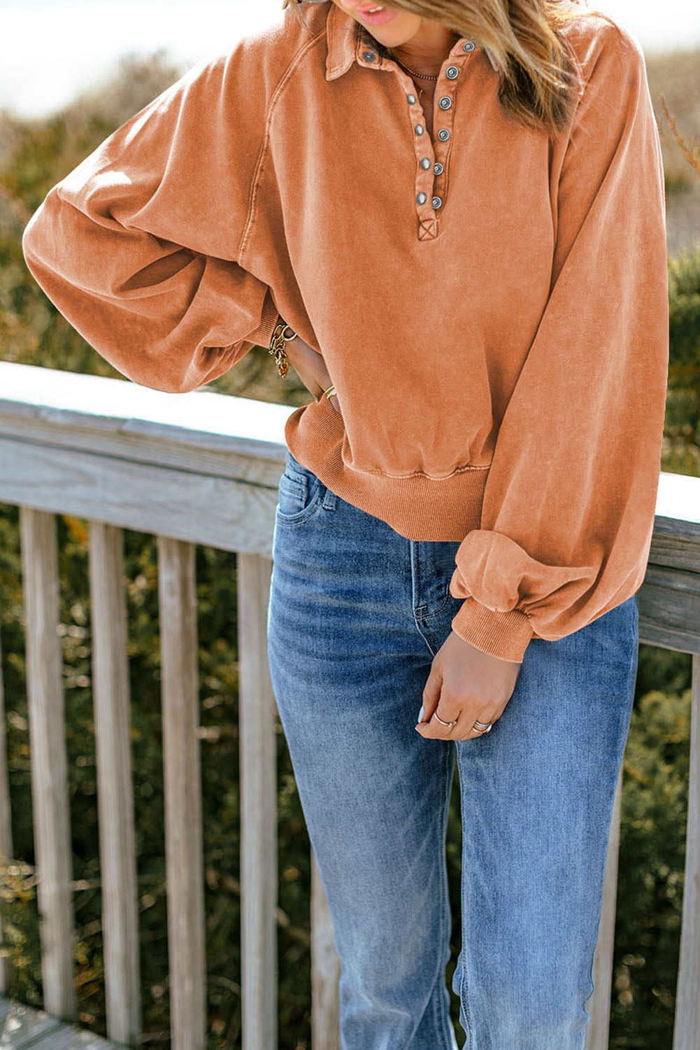 A person wearing an orange washed snap buttons lantern sleeve pullover sweatshirt and blue jeans stands with one hand on their hip, facing away from the camera, leaning slightly on a wooden rail outdoors.