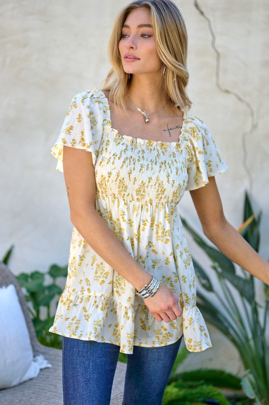 A woman stands outdoors donning a Floral Printed V-Neck Ruffle Top paired with jeans, set against a neutral background, with plants in the foreground, highlighting her fashion-forward wardrobe.